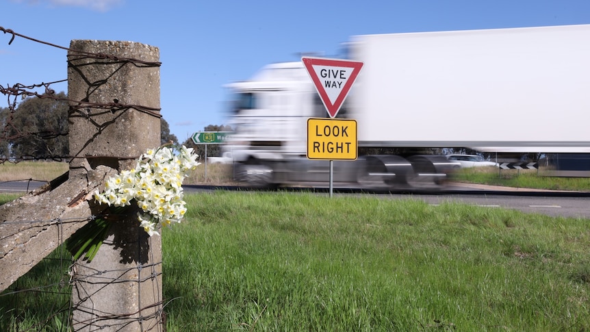 Flowers placed on a wooden post, grass patch ahead, a white truck whizzes past a give way/look right sign.