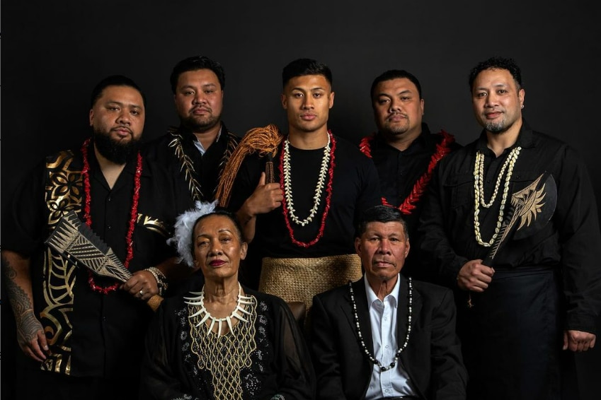 An older Samoan couple are seated in front, while the five brothers stand behind them, three carrying cultural tools.