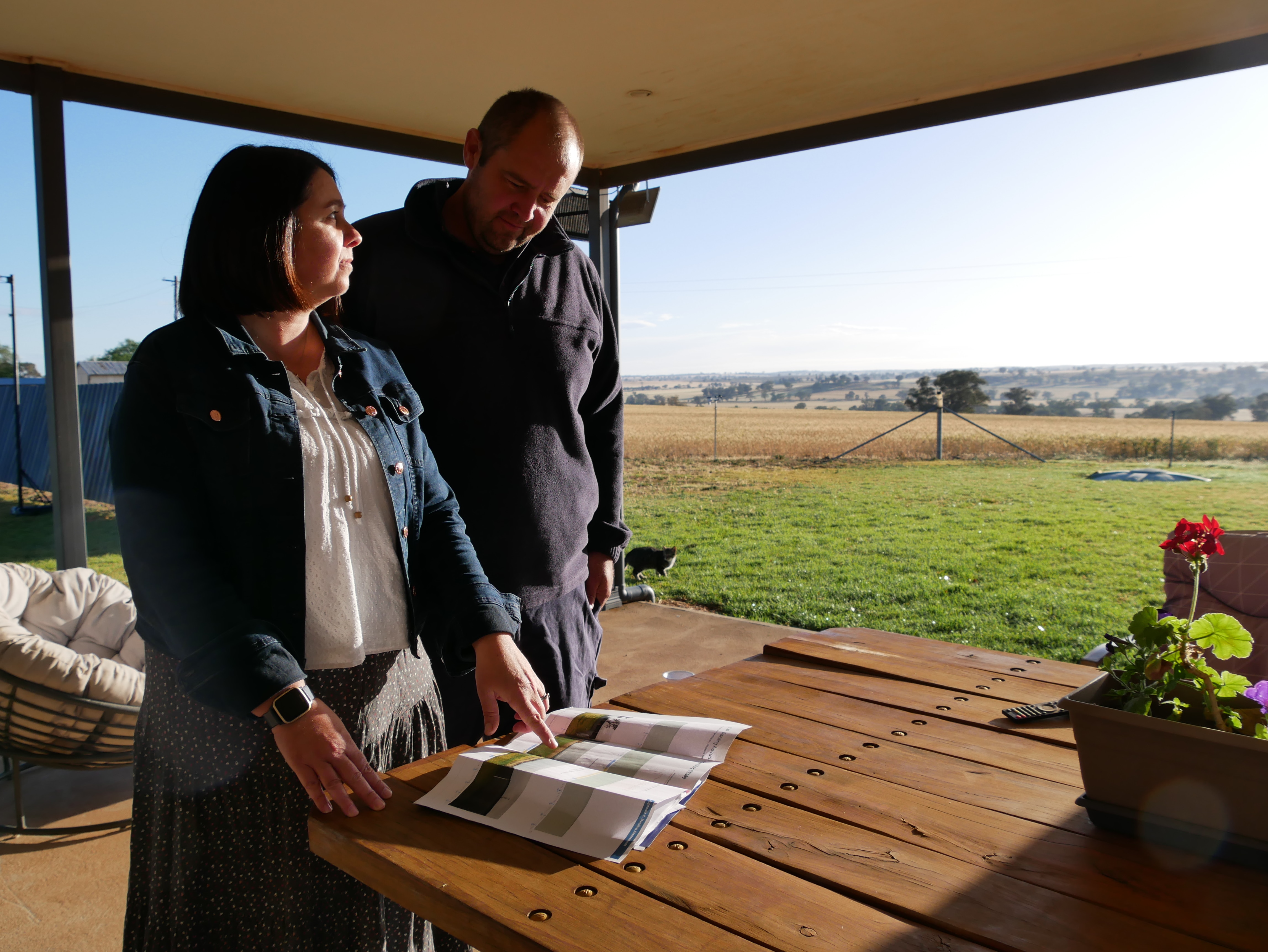A couple standing looking at plans on a back patio, with country side in the background.
