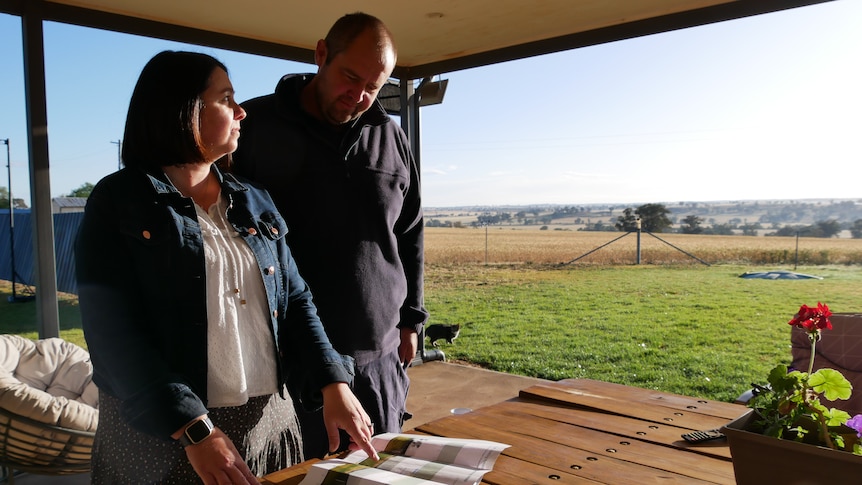 A couple standing looking at plans on a back patio, with country side in the background.