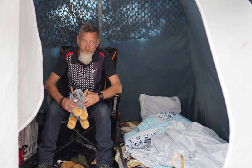 A man sits in a camping chair within a tent holding a soft toy doy, smiling at the camera.