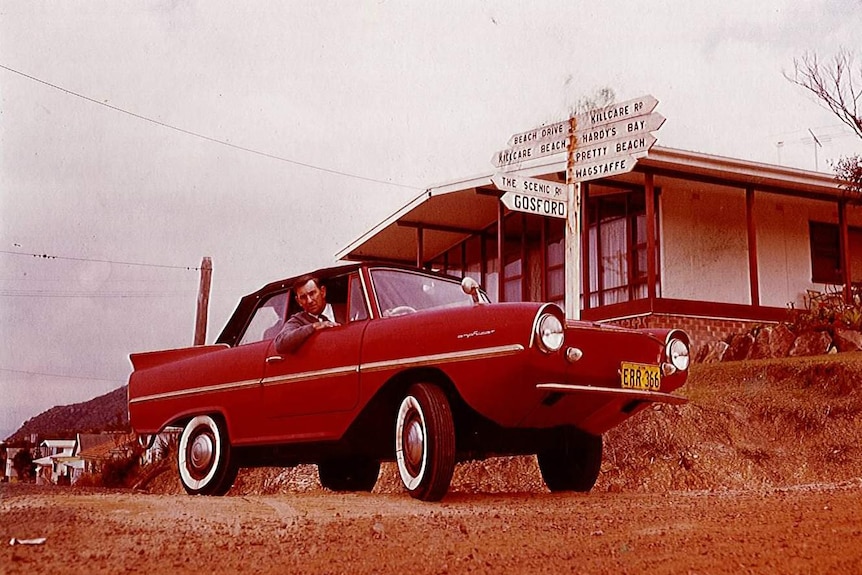 A man sitting inside a red car with his head outside the window looking at the camera. A sign points to Gosford and Killcare.