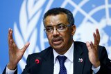 Tedros Adhanom Ghebreyesus sits at a microphone and gestures with his hands.
