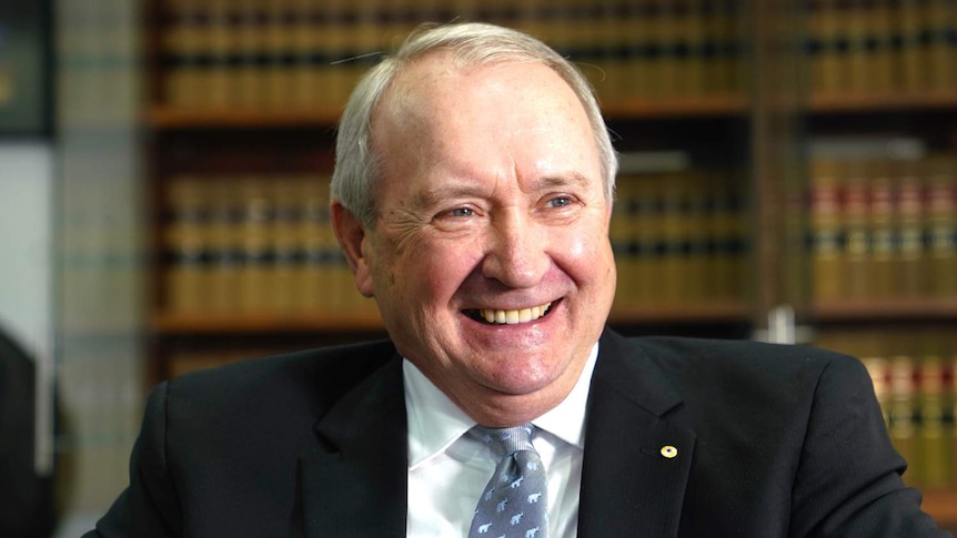 A man smiles broadly as he stands in front of shelves full of leather-bound books.