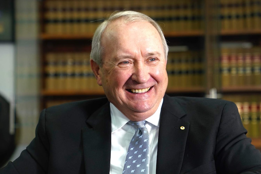 A man smiles broadly as he stands in front of shelves full of leather-bound books.