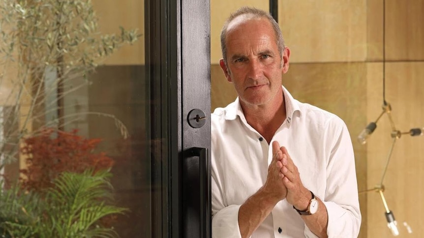 Kevin stands by a glass sliding door, rubbing his hands together and looking at the camera with a stern expression.