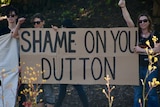 Three women stand near bushes with a sign reading "Shame on you Dutton"