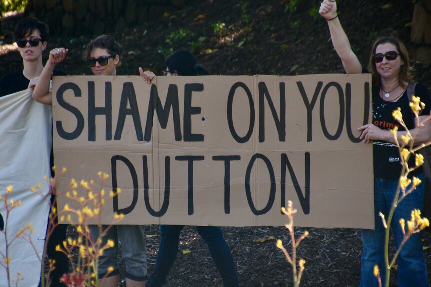 Three women stand near bushes with a sign reading "Shame on you Dutton"
