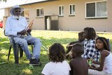 Kowanyama elder Colin Lawrence Snr sits in chair clapping two boomerangs with children sitting and watching in front of him.