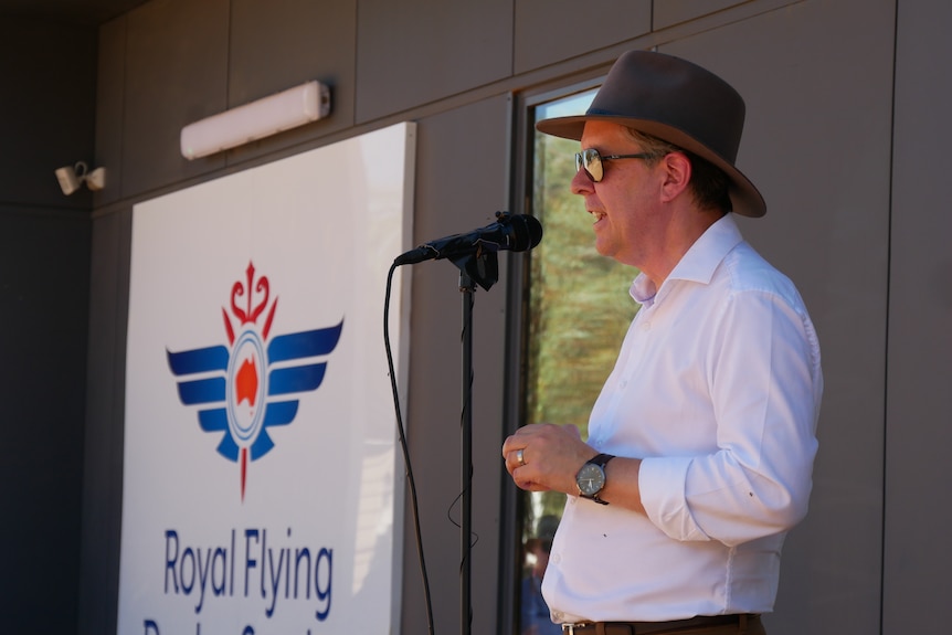 A man wearing an akubra hat, aviator sunglasses and a white shirt stands in front of a microphone. 