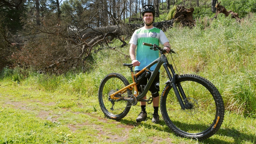 A white man with a helmet on stands with a yellow mountain bike on a nature path in front of a fallen, burnt pine tree.