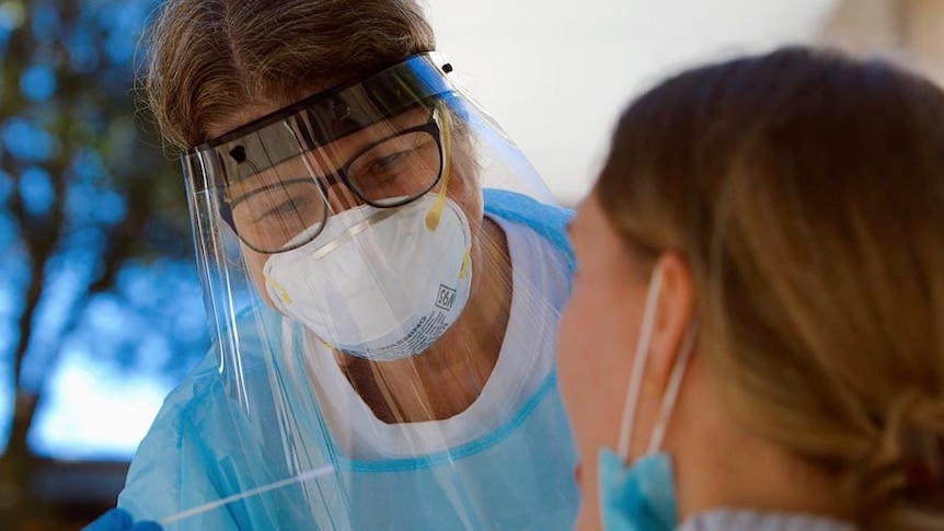 A nurse in PPE gear including clear full face mask and gloves holds a test swab beside a patient