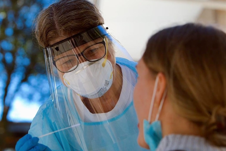A nurse in PPE gear including clear full face mask and gloves holds a test swab beside a patient