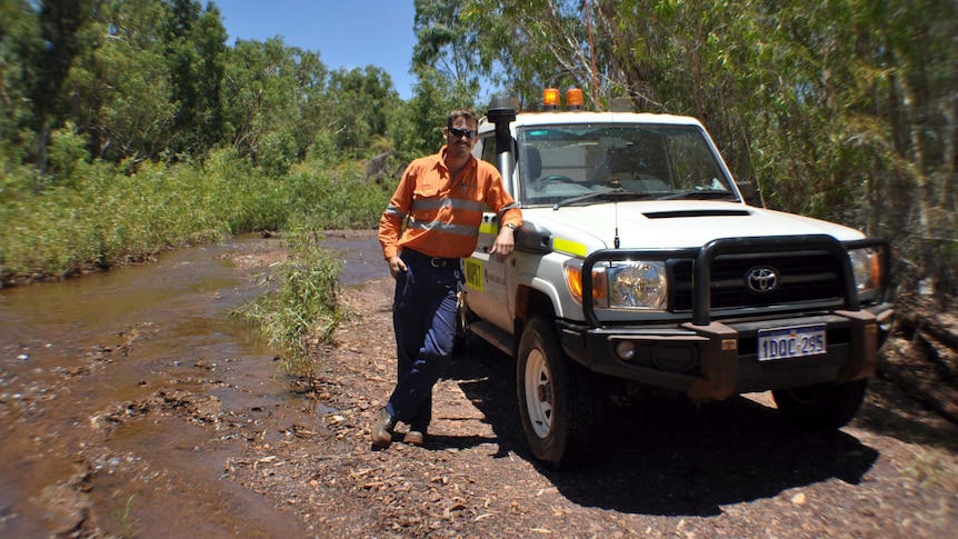 A man in a high vis shirt leans against a 4WD vehicle, next to a river.