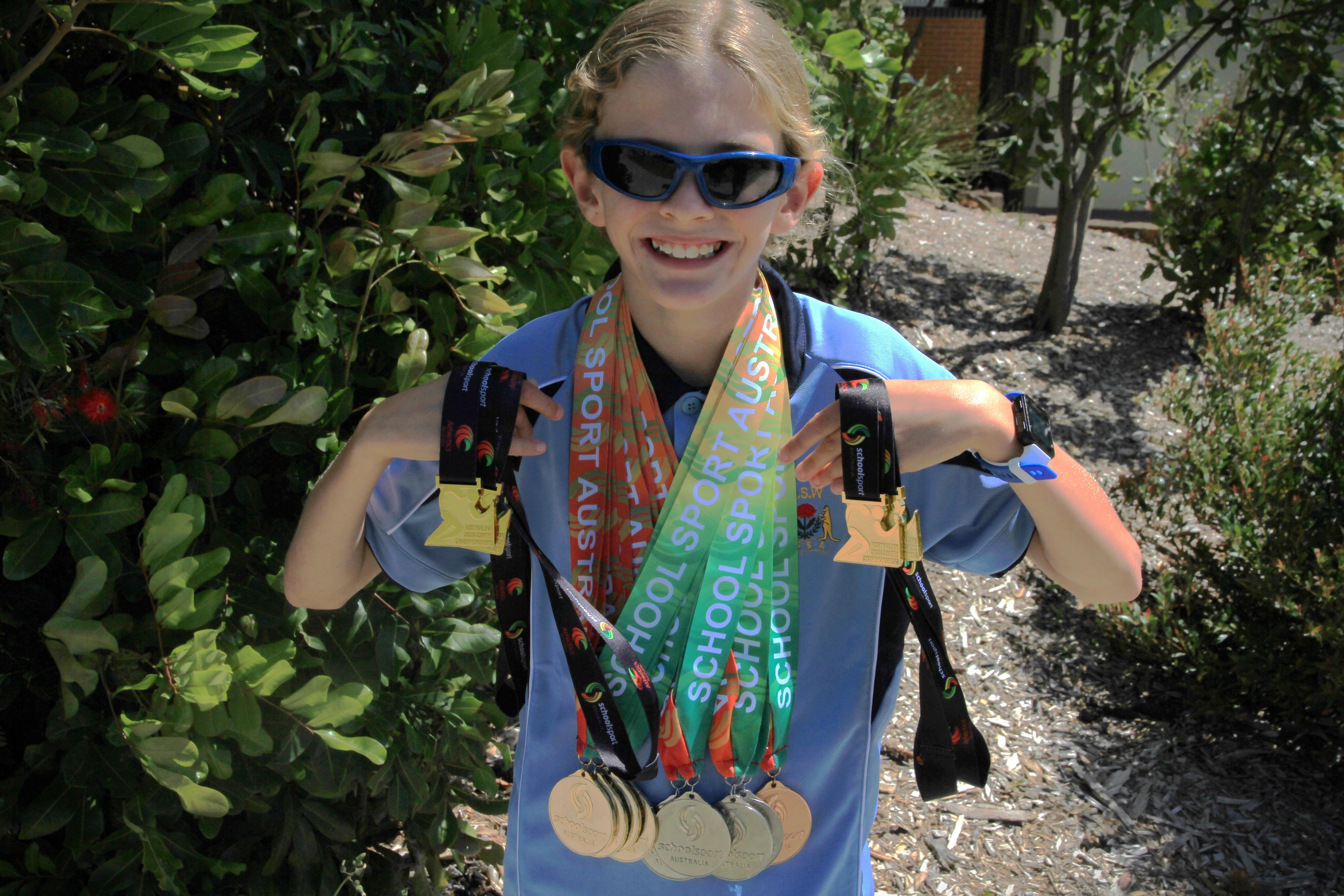 Blonde haired boy smiling, wearing dark sunglasses, coloured medals around his neck, blue uniform