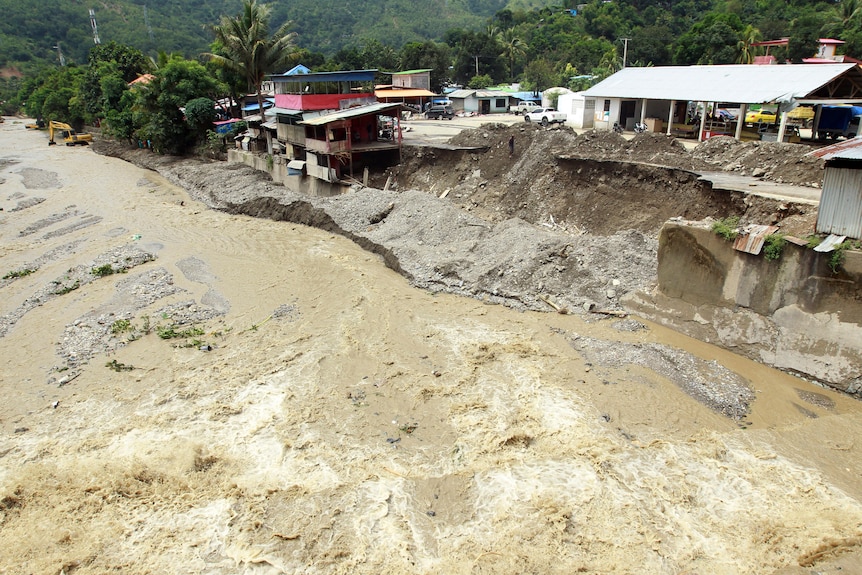 An aerial shot shows murky floodwaters surging past a large retaining wall that has collapsed. 