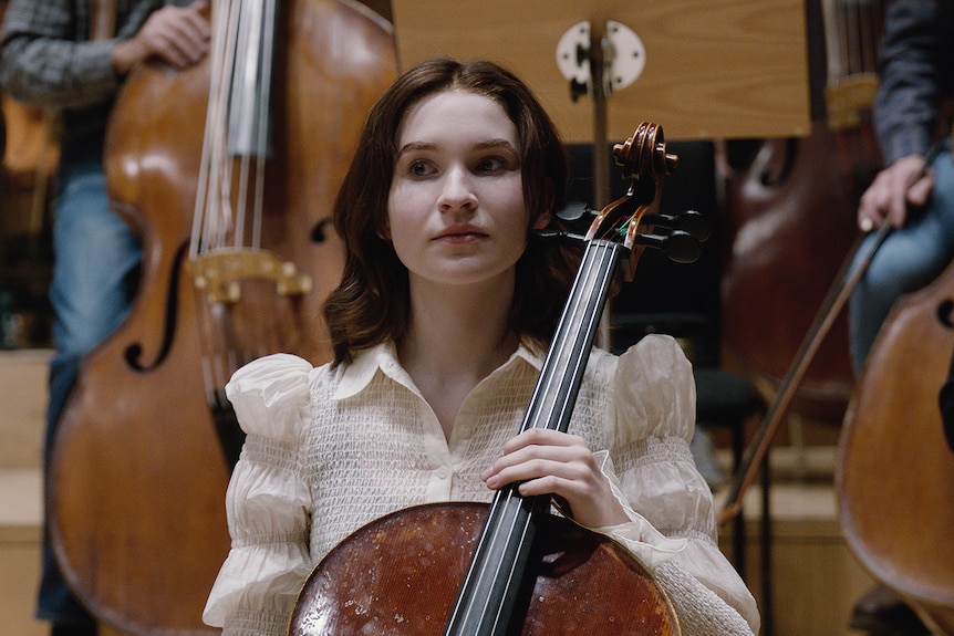 A brown-haired young woman sits with her back straight, holding the neck of a cello. Other cellists can be seen behind her.