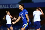 A female soccer player wearing blue celebrates after scoring a goal against a team in white