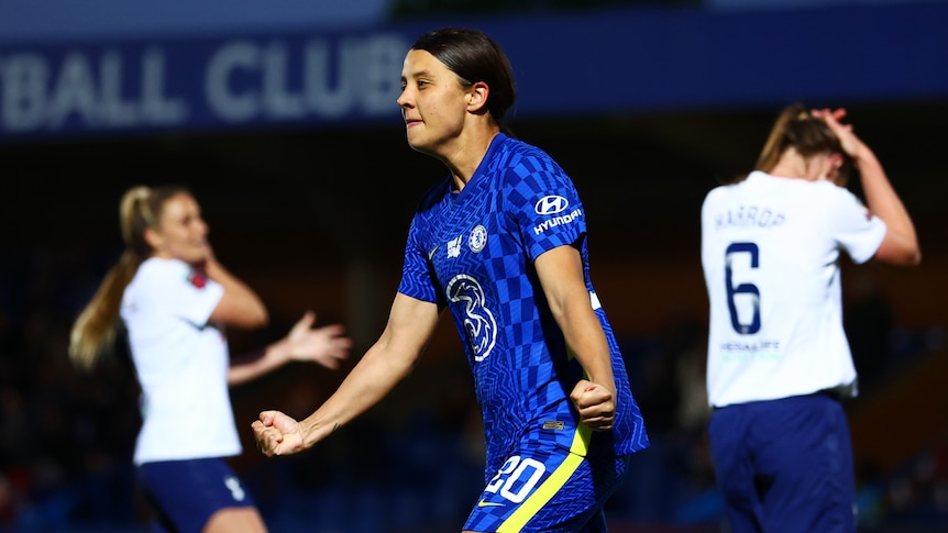 A female soccer player wearing blue celebrates after scoring a goal against a team in white