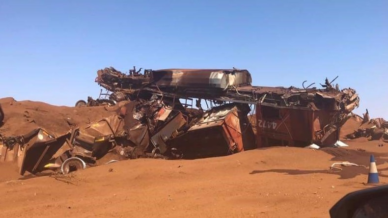 The mangled wreckage of an iron ore train sits on red dirt under a blue sky.