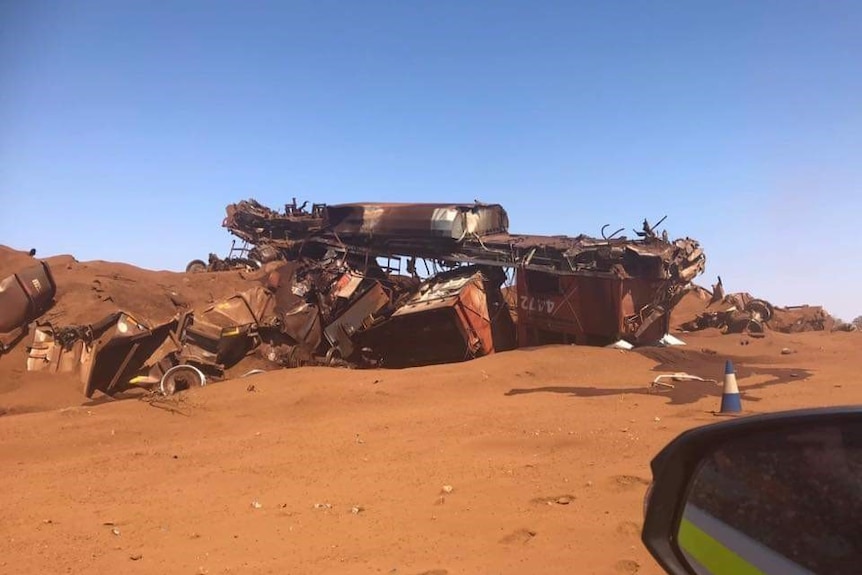 The mangled wreckage of an iron ore train sits on red dirt under a blue sky.