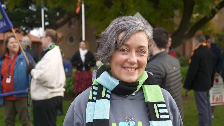 A woman with short grey hair, wears a green, black and blue scarf, holding a placard, smiling widely as people mingle behind.