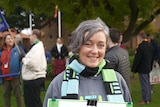 A woman with short grey hair, wears a green, black and blue scarf, holding a placard, smiling widely as people mingle behind.