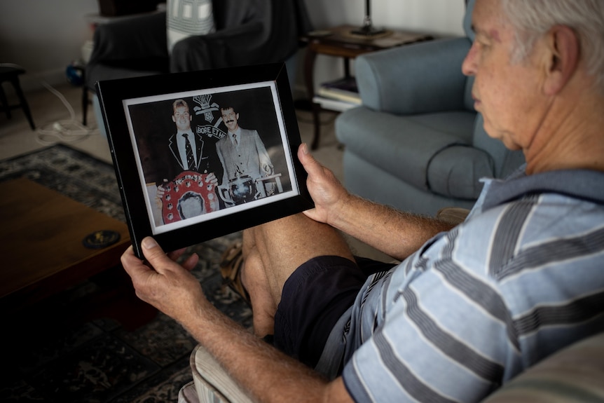 An older man looks at a photo of himself with a younger man from the 1980s.