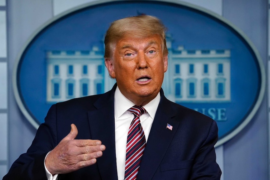 A man in a navy suit gestures with his right hand as he speaks while standing in front of a White House sign.