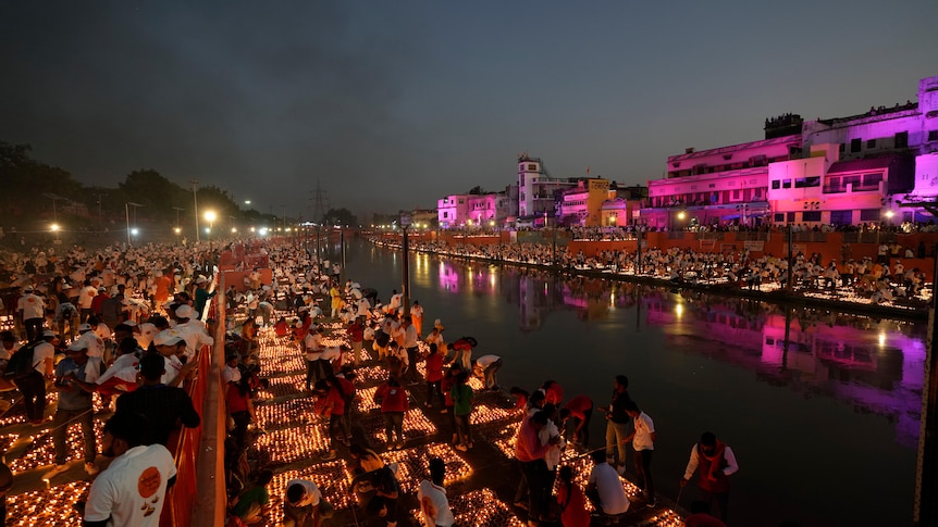 People light dozens of squares of lamps on the banks of a river. On the other side are more lamps and buildings lit up.