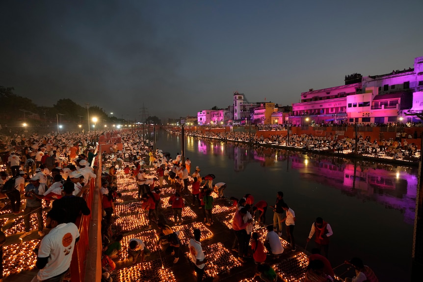 People light dozens of squares of lamps on the banks of a river. On the other side are more lamps and buildings lit up.