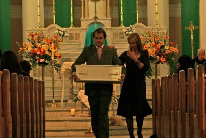 Alison and David Homer walking a small white casket down an aisle in a church.