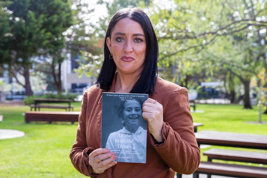 Author Beth Doherty holding a book she has written on the Catholic Church, with trees in background.