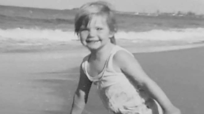 Toddler Cheryl Grimmer in a black-and-white photo, smiling at the shoreline of a beach.