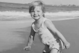A black-and-white photo of a smiling, fair-haired toddler playing near the tideline on a beach.