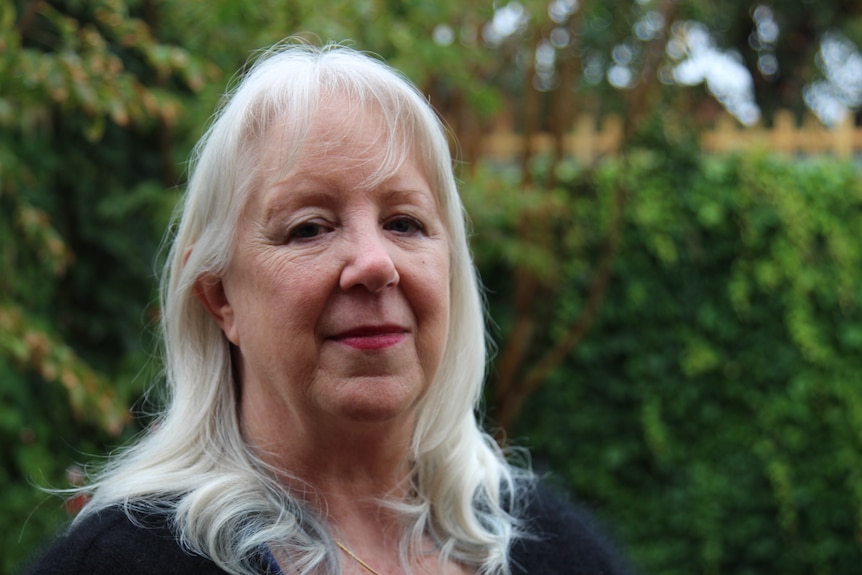 Close-up of Donna Ward, with shoulder-length blonde hair, and blurred greenery behind her, smiling slightly.