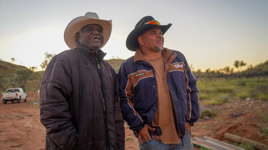 Two men, both smiling, look off-camera. They are wearing cowboy hats and are in the outback.
