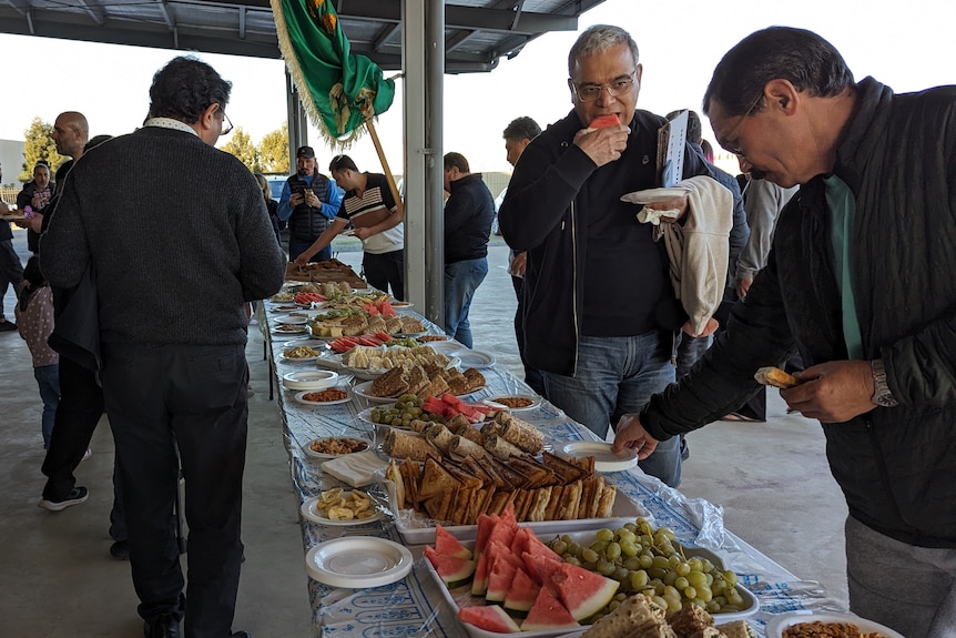 People eat some of the snacks, long table laden with food, including watermelon, grapes, sky, an awning over the table. 