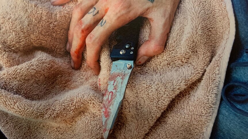 A hand with tattoos holding a bloodied knife sitting on a towel