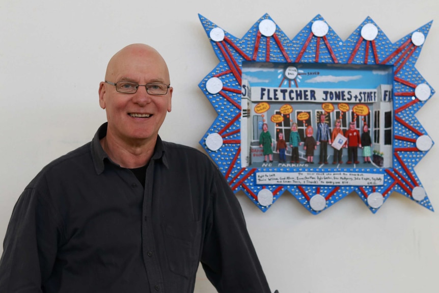 Artist Glenn Morgan stands next to a wooden relief work picturing Warrnambool locals