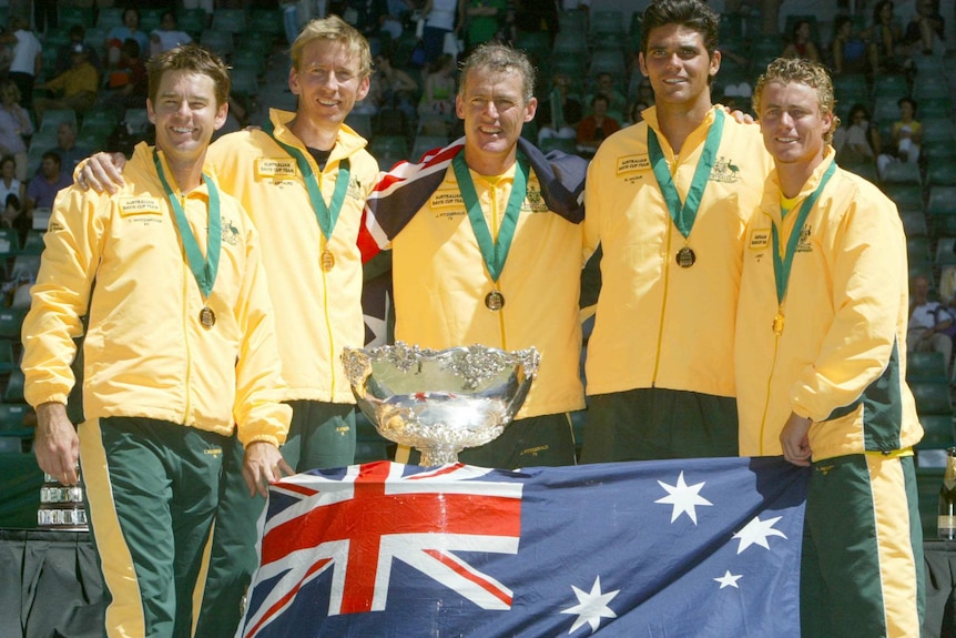 A tennis team stands together smiling with the Davis Cup trophy and an Australian flag.