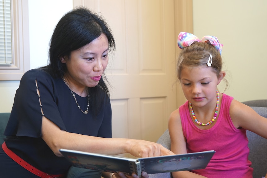 A middle aged lady aits beside a young girl and reads to her