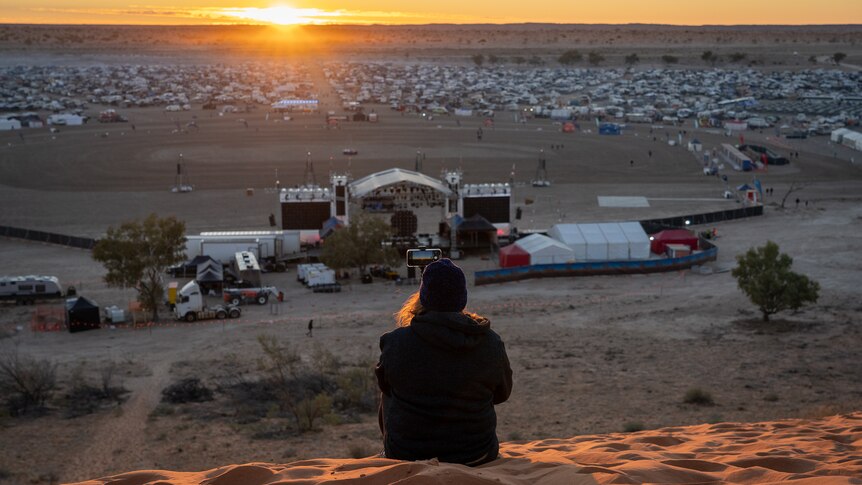 A person wearing a beanie sits on a hill of red dust overlooking a stage.