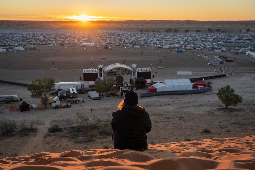A person wearing a beanie sits on a hill of red dust overlooking a stage.