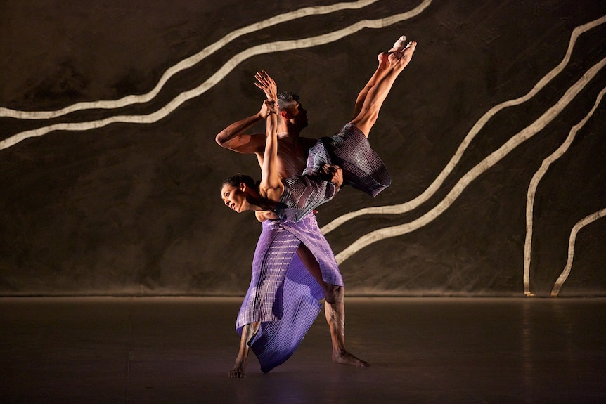 First Nations male dancer lifts First Nations female dancer - her feet are pointed and her arms stretched back like a bird.