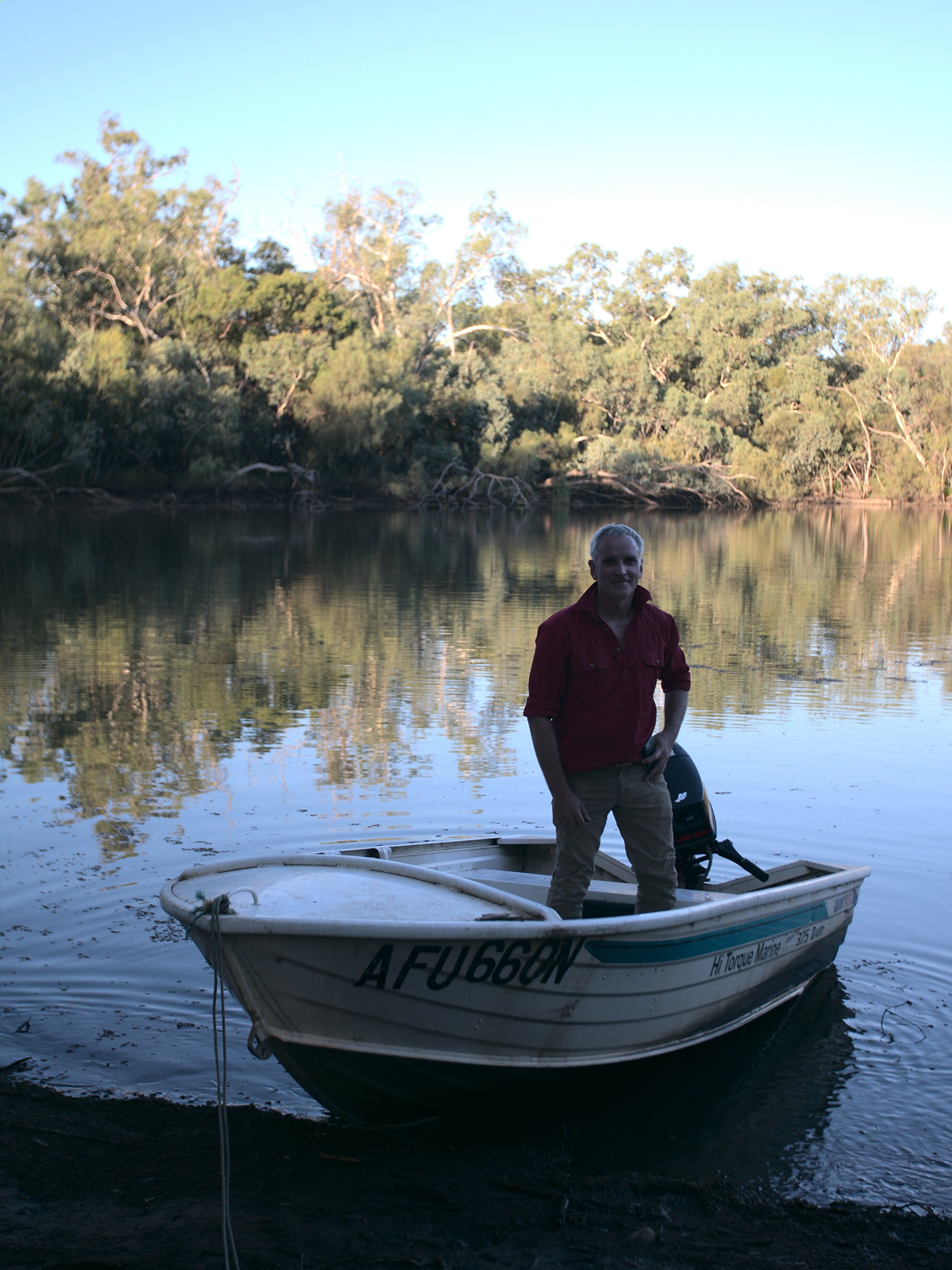 A man in a red shirt stands in a tinny just off the banks of a river.