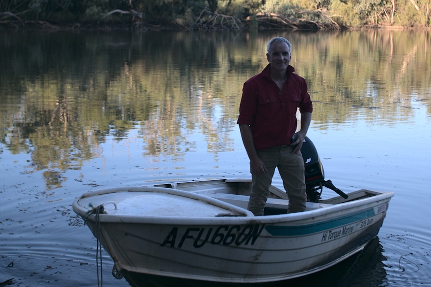 A man in a red shirt stands in a rowboat right by a river.