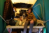 man with black beanie, brown coat and glasses sits on bed on flatbed ute with pet dog