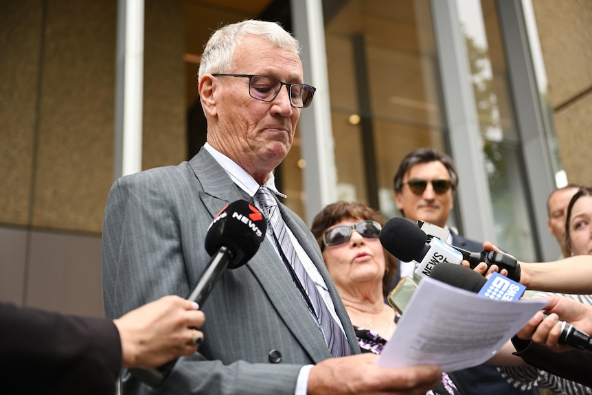 A man wearing glasses addresses reporters