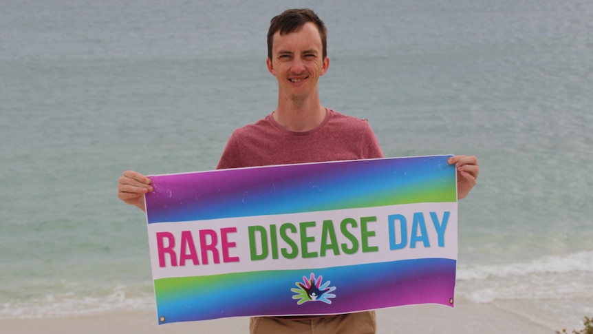 Andrew is holding a multi-coloured banner that says Rare Disease Day. Andrew has short dark hair and the ocean is behind him.